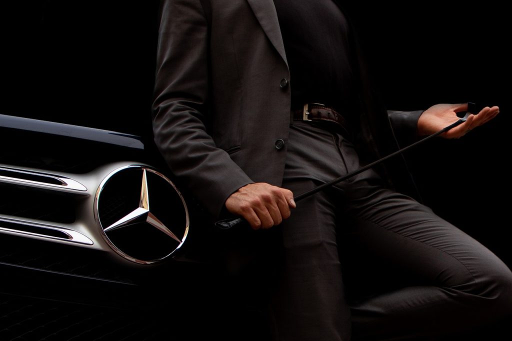 Man wearing gray suit leaning against a black Mercedes Benz car
